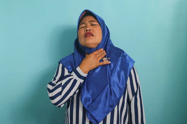 青いヒジャーブとストライプのシャツを着た中年のアジア人女性は 青い背景に立っている間 喉の痛みに苦しんでいます — ストック写真