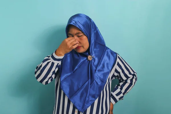 青のヒジャーブとストライプのシャツを着た中年のアジア人女性の肖像画は 彼女の鼻に触れると 青い背景に立って 片頭痛 または病気に苦しんでいることを示しています — ストック写真