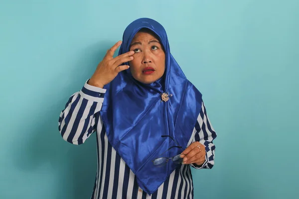 青いヒジャーブとストライプのシャツを着たアジア系の中年女性は 目の痛みに苦しみ 結膜炎のためにかゆみを感じた後 彼女の手を使って彼女の目をこすります 青い背景に隔離されました — ストック写真