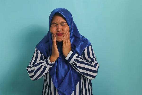 青のヒジャーブとストライプのシャツを着た中年のアジア人女性は 青い背景を背景に立ちながら厳しい歯痛に苦しんでいます — ストック写真
