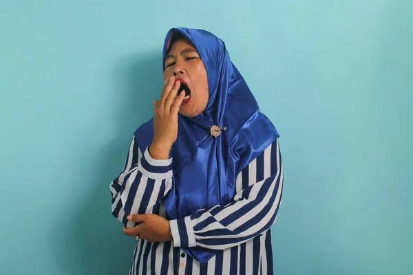 眠いアジアの女性は 青いヒジャーブとストライプのシャツを着て あくびをし 青の背景に立って 退屈と疲れて見て口をカバーしています 朝の疲労 — ストック写真
