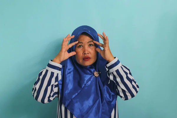 青いヒジャーブとストライプのシャツを着たアジア系の中年女性は 青い背景を背景に立ちながら 指で目を開けようとし 朝の疲労から退屈し疲れているように感じます — ストック写真