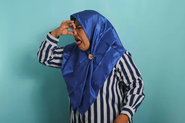 A middle-aged Asian woman, in a blue hijab, holds her breath and pinches her nose with her fingers, reacting to a stinky and disgusting, intolerable bad smell, while standing against a blue background