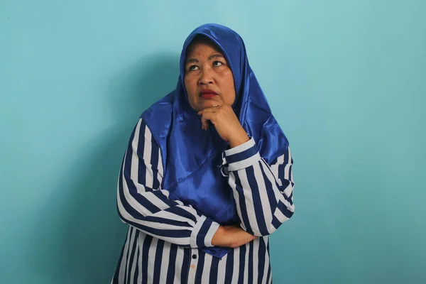 一位穿着蓝色头巾和条纹衬衫的忧心忡忡的亚洲中年妇女摸着下巴 站在蓝色的背景下 显得既困惑又忧虑 — 图库照片