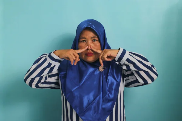 一名身穿蓝色头巾的亚洲中年妇女屏住呼吸 用手指捏住鼻子 对难闻难闻难忍的难闻气味做出反应 同时站在蓝色的背景下 — 图库照片