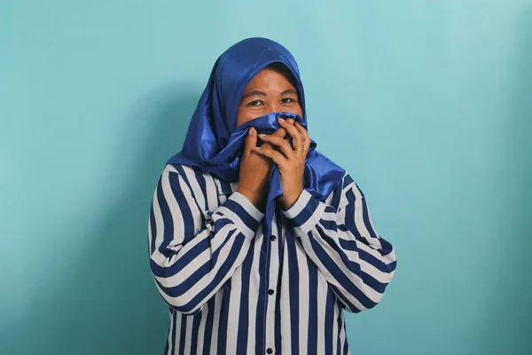 A middle-aged Asian woman, in a blue hijab, holds her breath and pinches her nose with her fingers, reacting to a stinky and disgusting, intolerable bad smell, while standing against a blue background