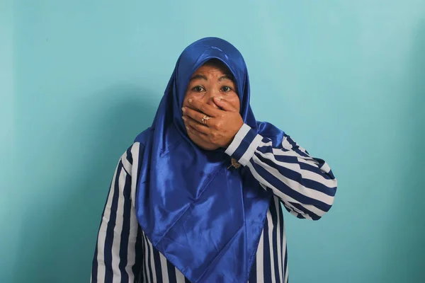 青いヒジャーブと口を覆うストライプのシャツを着たアジア系の中年女性がカメラを見て驚きました 青い背景に隔離された — ストック写真