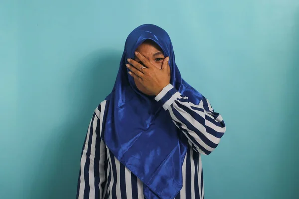 stock image Surprised middle-aged Asian woman in blue hijab and striped shirt covering mouth, staring at camera in astonishment. Isolated on blue background.