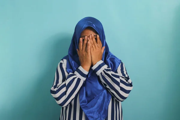 stock image A scared but curious middle-aged Asian woman in hijab hides her face, closes her eyes with her hands, and peeks through her fingers while watching a horror movie. Isolated on a blue background.