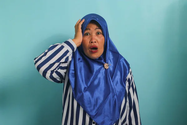 青いヒジャーブとストライプのシャツを着た怖がっている中年のアジア人女性は 青い背景を背景に立ちながら恐怖に飛び跳ねる — ストック写真
