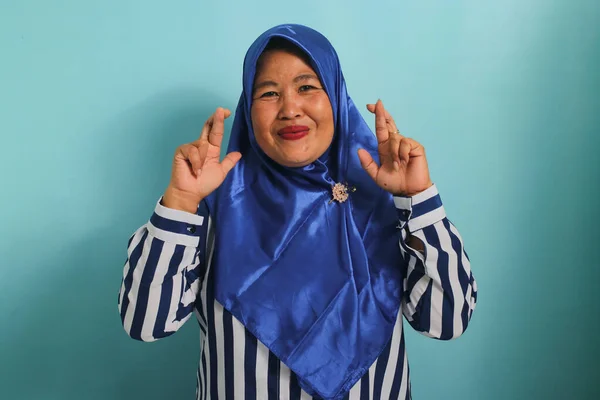 青のヒジャーブを身に着けている興奮した中年のアジアの女性は 希望と楽観的で 幸運のために彼女の指を渡り 願いを作り そして青の背景に立っている間笑顔です — ストック写真