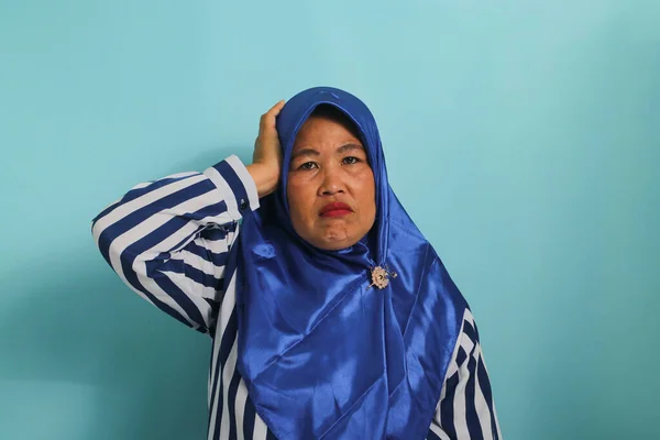 A troubled middle-aged Asian woman, wearing a blue hijab and a striped shirt, grabs her head and stares alarmed at the camera, feeling anxious while standing against a blue background