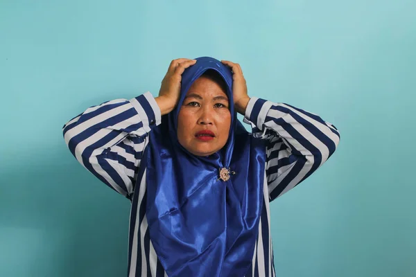 A troubled middle-aged Asian woman, wearing a blue hijab and a striped shirt, grabs her head and stares alarmed at the camera, feeling anxious while standing against a blue background