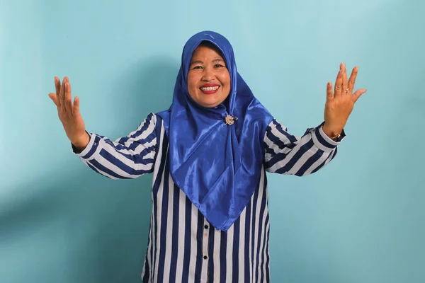 青いヒジャーブとストライプのシャツを着た興奮したアジアの女性は 青い背景に立って 開いている腕と大きな笑顔で皆を歓迎します — ストック写真