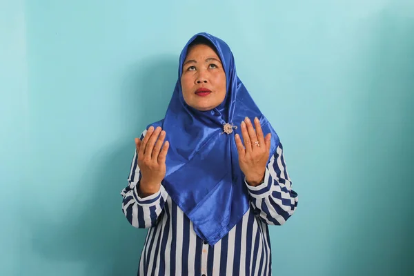 青いヒジャーブとストライプのシャツを着た中年のアジア人女性の肖像は 彼女の手を上げて祈りのジェスチャーを作っています 彼女は青い背景に隔離されている — ストック写真