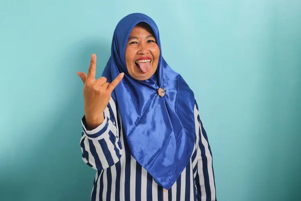 一位穿着蓝色头巾和条纹衬衫的快乐的中年亚洲女人 手拿着手势 露出她的舌头和对重金属的热爱 在蓝色的背景下显得孤立无援 — 图库照片