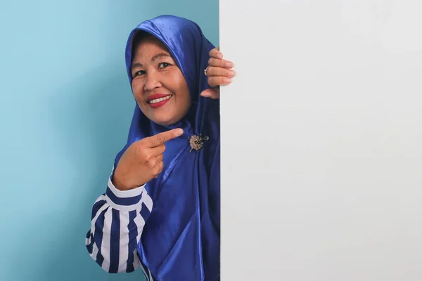 青のヒジャーブとストライプのシャツを着た興奮した中年のアジアの女性は 青い背景を背景に あなたのメッセージのための十分なスペースを提供し コピースペースと空白の白いバナー看板を保持し 指しています — ストック写真