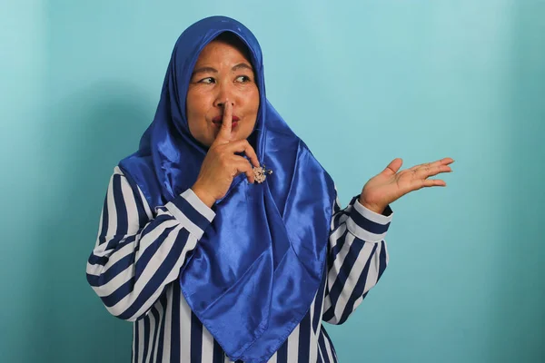 青いヒジャーブを纏った中年のアジア人女性が 余白に指を向けている 彼女は急いでいるか静かなジェスチャーをし 秘密の販売の申し出を伝え 青い背景に隔離されています — ストック写真