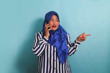 Mavi tesettürlü ve çizgili tişörtlü orta yaşlı Asyalı bir kadın telefonda konuşurken doğru telif alanını gösteriyor. Mavi bir arka planda izole edilmiş.
