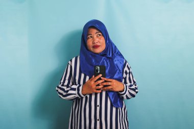 Mavi tesettürlü, çizgili tişörtlü, elinde cep telefonu olan, mavi arka planda izole edilmiş, orta yaşlı düşünceli, düşünceli bir Asyalı kadın.