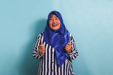 Mavi tesettürlü ve çizgili tişörtlü heyecanlı bir orta yaşlı Asyalı kadın elinde çatal ve kaşıkla yemek servisi beklerken acıkıyor. Mavi bir arka planda izole edilmiş.