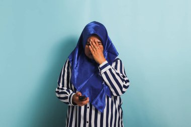 Mavi tesettürlü ve çizgili tişörtlü orta yaşlı endişeli bir Asyalı kadın, elinde bir televizyon kumandası tutarken stresli bir şekilde kafasına dokunuyor. Mavi bir arka planda izole edilmiş.