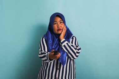 Mavi tesettürlü ve çizgili tişörtlü, orta yaşlı bir Asyalı sıkıcı bir televizyon programı izliyor ve uzaktan kumandayla kanal değiştiriyor. Mavi bir arka planda izole edilmiş..