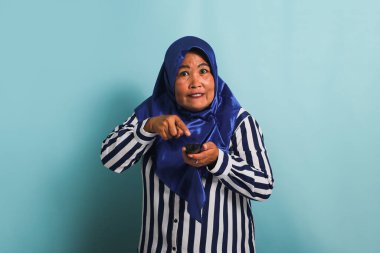 Mavi tesettürlü ve çizgili tişörtlü, sinirli, orta yaşlı Asyalı bir kadın televizyonda film izlerken kanal değiştirmeye çalışıyor. Mavi bir arka planda izole edilmiş.