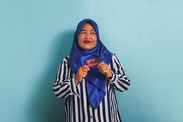青のヒジャーブとストライプのシャツを着て 青の背景に隔離されたオンラインビジネスを支払うためのクレジットカードを提示し 興奮した中年アジアの女性 — ストック写真