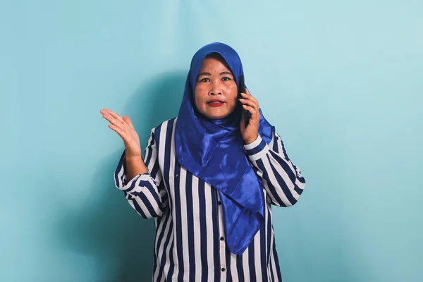 青のヒジャーブを着たアジア系の中年女性とストライプのシャツが電話をかけ 失望した怒りの表情で議論している 彼女は青い背景に隔離されている — ストック写真