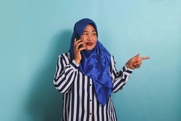 青いヒジャーブを着たアジア系の中年女性とストライプのシャツが 電話で話している間 右側のコピースペースを指しています 彼女は青い背景に隔離されている — ストック写真