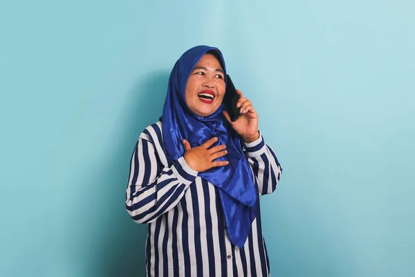 青いヒジャーブとストライプのシャツを着た興奮した中年のアジア人女性が 携帯電話で話している間に笑っている 彼女は青い背景に隔離されている — ストック写真