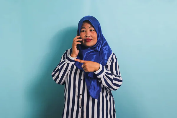 青いヒジャーブを着たアジア系の中年女性とストライプのシャツが電話で話している間に左側のコピースペースを指しています 彼女は青い背景に隔離されている — ストック写真