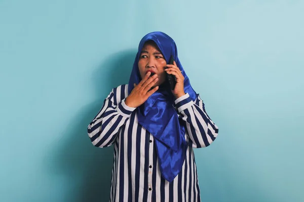一名身穿蓝色头巾和条纹衬衫的震惊的亚洲中年妇女带着惊讶的表情在电话里说着话 听到了一个重大新闻 她被蓝色背景隔离了 — 图库照片