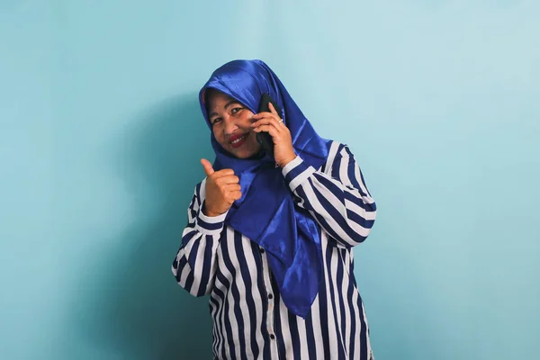 一位穿着蓝色头巾和条纹衬衫的快乐的亚洲中年女性一边打电话一边竖起大拇指 她被蓝色背景隔离了 — 图库照片