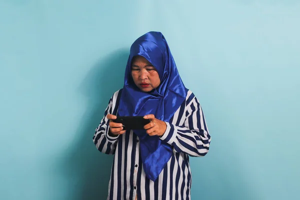 A bored middle-aged Asian woman in a blue hijab and a striped shirt is playing a game on her mobile phone, gesturing reluctantly, looking sad, and losing the game, isolated on a blue background.