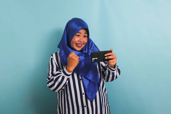 青のヒジャーブとストライプのシャツに興奮した中高年アジアの女性 携帯電話でゲームをプレイ 青の背景に隔離された — ストック写真