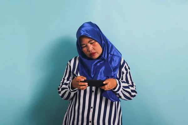 青のヒジャーブとストライプのシャツを着た退屈な中年のアジア人女性が 携帯電話でゲームをして しぶしぶジェスチャーをし 悲しい顔をし ゲームを失い 青の背景に隔離されています — ストック写真
