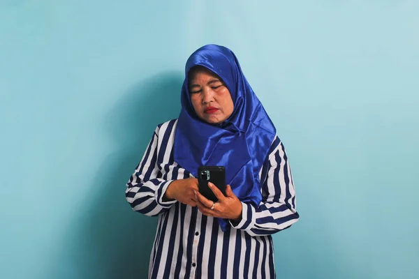 一位穿着蓝色头巾和条纹衬衫的无聊的亚洲中年女性拿着一部手机 看起来很无聊 不知如何是好 她被蓝色背景隔离了 — 图库照片