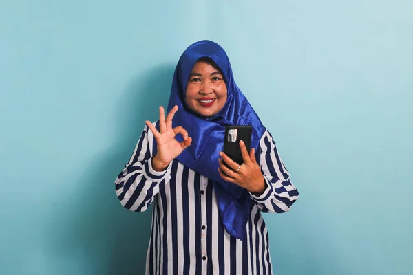 青いヒジャーブとストライプのシャツを着た興奮した中年アジアの女性が 承認サインやOkジェスチャーをしながら彼女の電話を握っています 彼女は青い背景に隔離されている — ストック写真