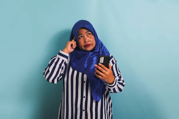 身穿蓝色头巾和条纹衬衫 手持手机 心事重重的亚洲中年女性 带着蓝色背景的蓝色背景 想着一个有趣的建议 — 图库照片