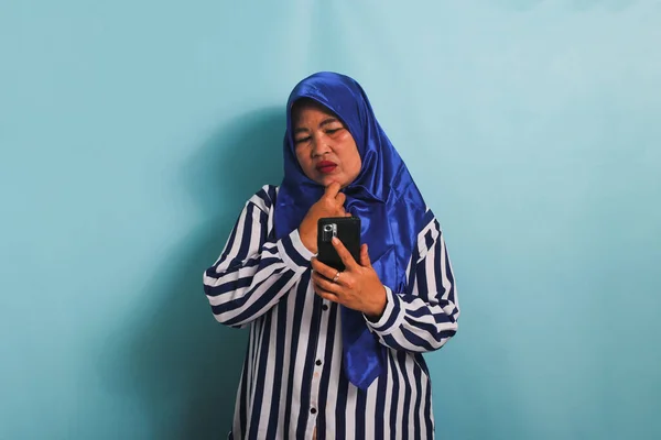 青のヒジャーブとストライプのシャツを着たアジア系の中年女性が携帯電話を持ち 興味深い提案を考え 青の背景に隔離された — ストック写真