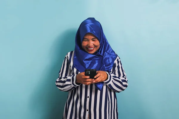 一名身穿蓝色头巾和条纹衬衫的兴奋的亚洲中年妇女拿着手机 脸上流露出热情的表情 她被蓝色背景隔离了 — 图库照片