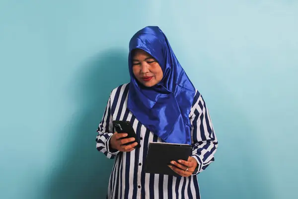 身穿蓝色头巾和条纹衬衫的中年妇女 手持一本书和一部手机 与蓝色背景隔离 — 图库照片