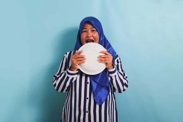 stock image A sweet and funny middle-aged Asian woman in a blue hijab and a striped shirt licks her plate, still hungry after eating. She is isolated on a blue background