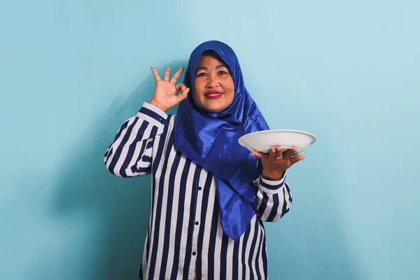 青いヒジャーブとストライプのシャツを着た中年のアジア人女性が 空の白いプレートや皿を持ちながらOkサインを示しています 彼女は青い背景に隔離されている — ストック写真