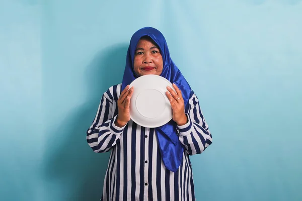 青いヒジャーブと縞模様のシャツを着た不幸な中年のアジア人女性は 食事中や断食中に空腹を感じる空の白いプレートを保持しています 彼女は青い背景に隔離されている — ストック写真