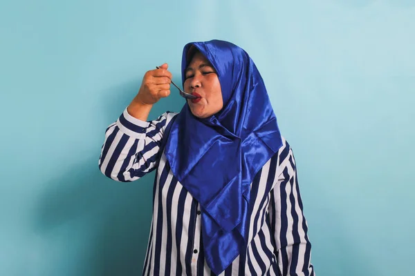 一名身穿蓝色头巾和条纹衬衫的亚洲中年妇女 举着一只金属勺子 闭着眼睛 正在品尝食物 她被蓝色背景隔离了 — 图库照片