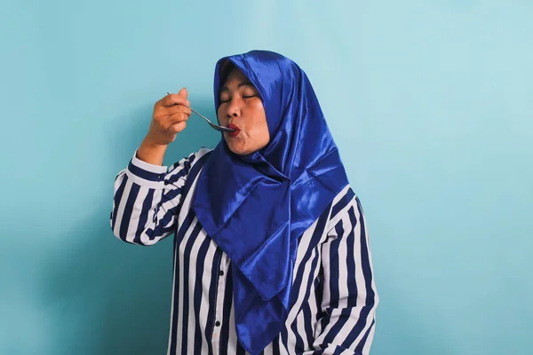 一名身穿蓝色头巾和条纹衬衫的亚洲中年妇女 举着一只金属勺子 闭着眼睛 正在品尝食物 她被蓝色背景隔离了 — 图库照片