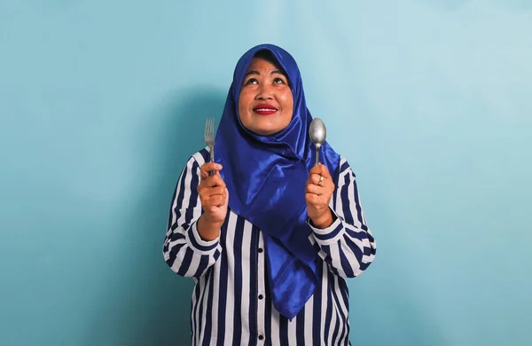 一名身穿蓝色头巾和条纹衬衫的兴奋的亚洲中年妇女用勺子和叉子指向上方的彩色空间 她被蓝色背景隔离了 — 图库照片